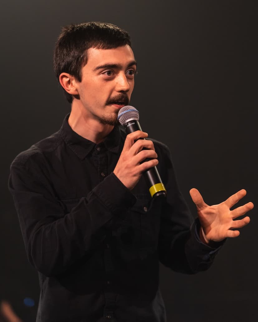 Cal is hosting a show. He's wearing a black shirt and smiling as he speaks into a microphone.